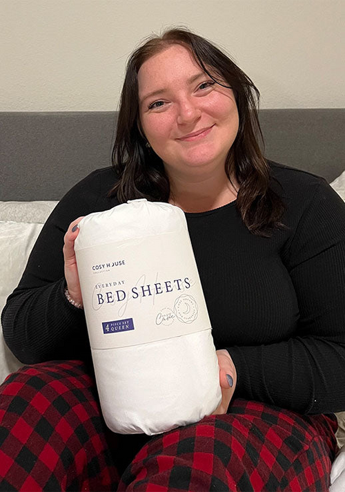 A smiling female customer is holding a packaged set of Cosy House Collection’s Everyday Bed sheets.