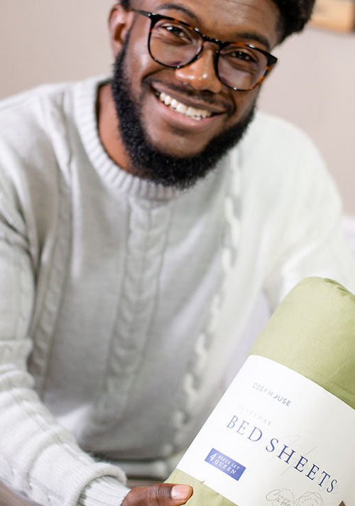 A smiling male customer is holding a packaged set of Cosy House Collection’s Everyday Bed sheets.