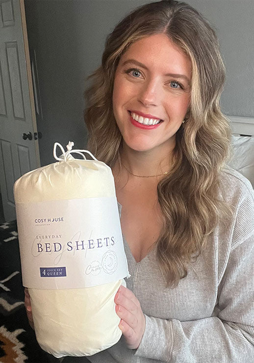 A happy female customer is holding a packaged set of Cosy House Collection’s Everyday Bed sheets.