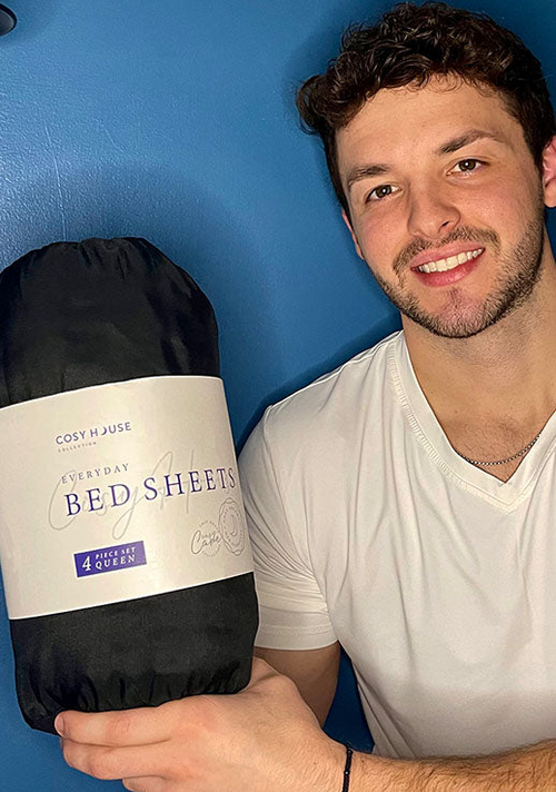 A happy male customer is holding a packaged set of Cosy House Collection’s Everyday Bed sheets.
