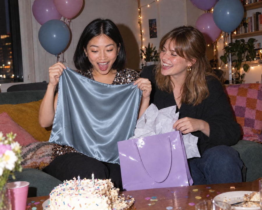 Two friends at a birthday party, one holding up an ash-blue silk pillowcase from a lilac gift bag